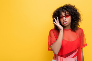 trendy african american woman in red top adjusting sunglasses isolated on yellow.