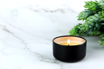 aroma scented candle in black tin cup lighting on the white marble table with plants for relax...