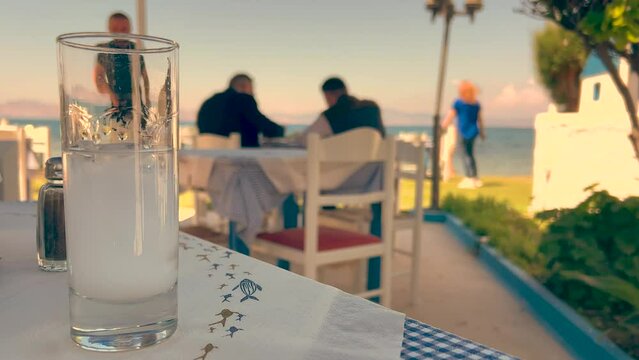 Filled Raki, Saki or Ouzo spirit glass on table, close-up. Milky alcoholic liqueur, selective focus. Unrecognizable people moving in background. 4K resolution for advertisement, video banner