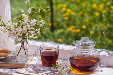 Hot tea in glass teapot and cup on windowsill at home