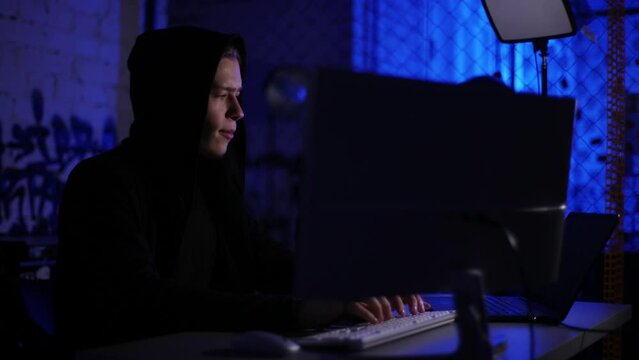 Portrait of confident hacker man in black sweatshirt using laptop and desktop computer to hack network system from dark room with blue neon lights. Hacker typing program code committing cybercrime.