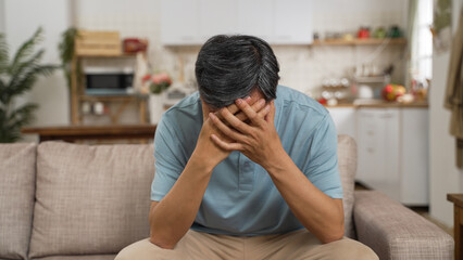 Closeup view of ill Asian senior man covering face and propping head with both hands while suffering dizziness on sofa in the living room at home.