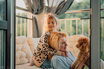 happy mother and daughter are sitting at home on the balcony on a light sofa with a pet: a poodle dog on a warm sunny evening at sunset at the golden hour