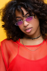 brunette african american woman in necklaces and trendy sunglasses smiling at camera isolated on yellow.