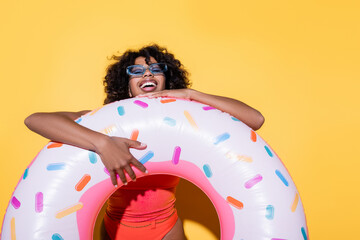 excited african american woman in trendy sunglasses laughing near swim ring on yellow background.