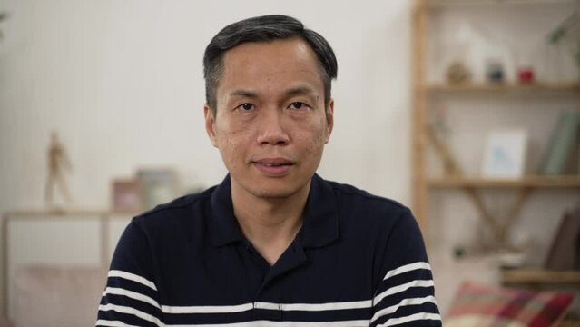 half length portrait of a grey haired Asian male pensioner in casual wear looking at camera with serious face expression on home background