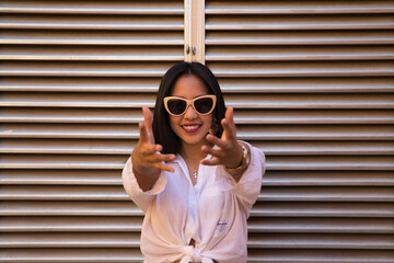 young, beautiful, dark-haired South American woman, wearing a white shirt and sunglasses, has a silver-coloured air outlet in the background. The woman raises her hands in front of her.