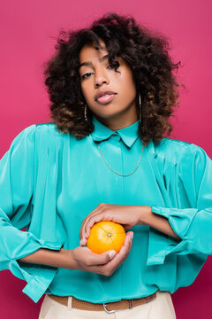 african american woman in trendy cyan blouse holding ripe orange isolated on pink.