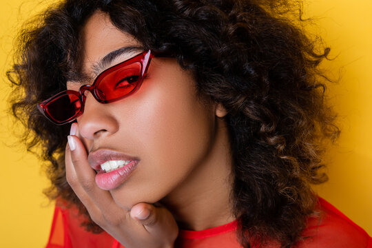 portrait of african american woman in red sunglasses touching face isolated on yellow.