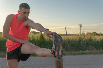 Young runner man doing leg stretches on a wooden pole. Side view of a sporty boy who has one leg raised on a piece of wood to stretch the muscles of the lower body after a great and demanding effort. - obrazy, fototapety, plakaty