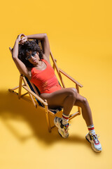 high angle view of african american woman in beachwear relaxing in deck chair on yellow background.