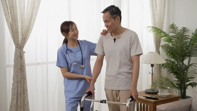 cheerful asian older man under recovery process is talking happily with female nursing aide by window in a bright home interior. he stands with the support of walker