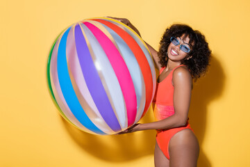 joyful african american woman in swimsuit and trendy sunglasses holding striped inflatable ball on yellow background.