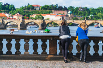 Two young women near the stone railings of the bridge in Prague, Czech Republic, admiring the view of the Vltava river and the old town on a sunny day, Europe tourist travel, back view