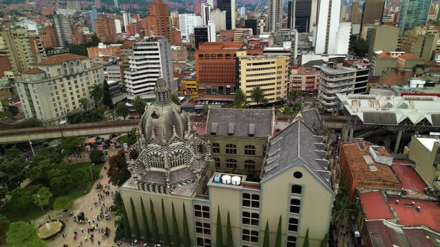 The Catholic Basilica of Medellin stands by a park through which people pass. Surrounded by urban architecture, passing cars and train tracks.