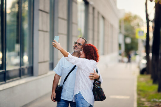 Cute middle-aged european couple with smart phone taking pictures, taking selfies while walking on the city street, summer walks and travel
