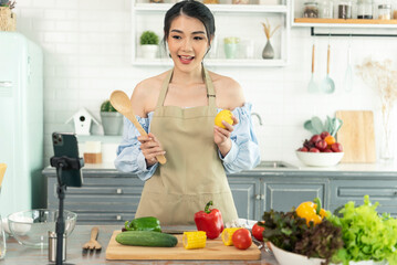 Asian woman food blogger cooking salad in front of smartphone camera while recording vlog video and live streaming at home in kitchen.
