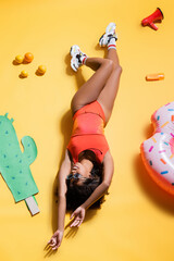 top view of african american woman in trendy beachwear near paper cut cactus, swim ring and fruits on yellow.