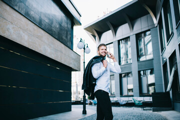 Young cheerful man talking on smartphone beside modern buildings on street