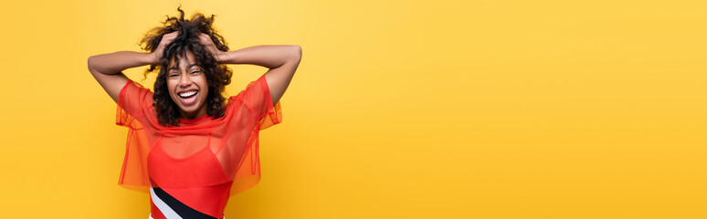 cheerful african american woman in red summer top holding wavy hair isolated on yellow, banner.