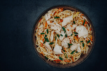 Fried Chinese noodles with chicken, carrots and green onions in a bowl with a dark background photographed from above