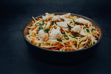 Fried Chinese noodles with chicken, carrots and green onions in a bowl with a dark background