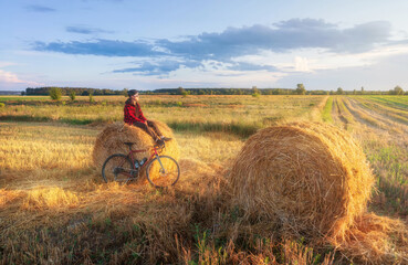 Obraz premium Cyclist sits on a haystack after a bike ride at sunset. Active lifestyle and travel concept.