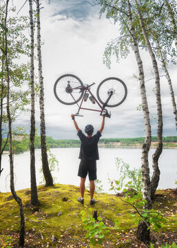 Active young guy holding a bicycle over his head on the background of a lake in the woods. Sports and travel.