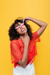 african american woman touching chin and wavy hair while posing in sunglasses isolated on yellow.