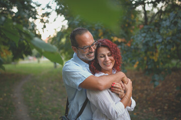 Fototapeta premium red-haired pretty woman hugs slender man with glasses. Cute middle aged european couple hugging in the park