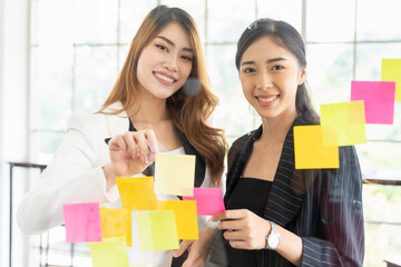 Group of business Asian women meeting at the office and use post it notes on glass wall. Business Asian women using sticky note brainstorming and share ideas on glass wall.