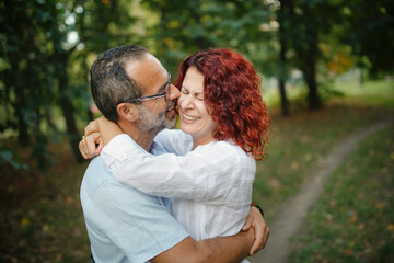 slender man with beard kisses and hugs pretty red-haired curly woman. Cute middle aged european couple hugging in the park
