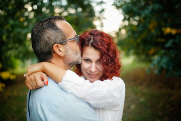 slender man with beard kisses and hugs pretty red-haired curly woman. Cute middle aged european couple hugging in the park