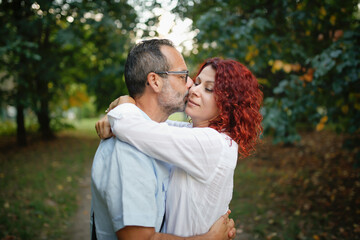 Fototapeta premium slender man with beard kisses and hugs pretty red-haired curly woman. Cute middle aged european couple hugging in the park