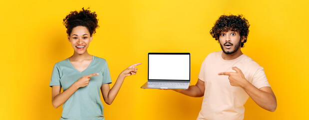 African american girl and arabian or indian guy, hold an open laptop with blank white mock-up screen for advertisement or presentation, stand on isolated orange background, look at camera, smile