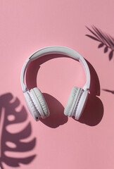 Wireless white headphones with tropical leaves shadows background on the pink close up. Listening to summer music concept	