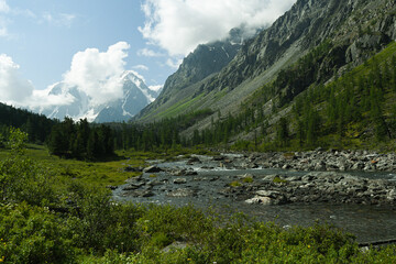 Fototapeta na wymiar Mountain stream during spring flood, water breaks against rocks. Rivulet in mountainous area, rock ridge on horizon. Glaciers and snow on hillsides. Clouds over riverbed.