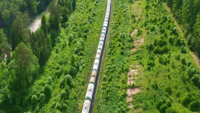 Top, aerial view of long freight train that moving along green forest in summer. Locomotive, tank wagons ride on rails carrying liquid or bulk cargo. Railroad transportation, delivery taken by drone