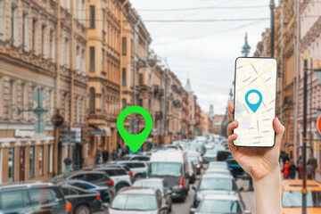 A female's hand holds a smartphone with an online map app. In the background is a city street with traffic jam, with a green location icon.Concept of online navigation and GPS