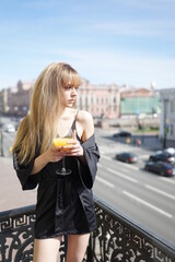 In the afternoon, in the light of the sun, a touching girl in black pajamas enjoys a glass of orange juice. She's good on the balcony.
