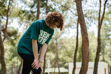 Naklejka premium Side view of young redhead runner female wearing sportive clothes standing on city park, outdoors touching injured knee with hands. Injured leg, healthy lifestyle and sport concept.