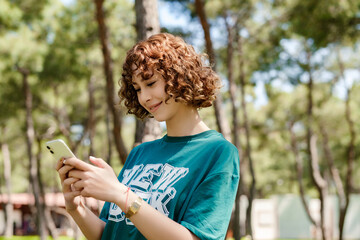 Young beautiful redhead woman wearing green t-shirt and standing on city park, outdoors looking at the phone screen and using phone. Messaging with friends, watching video or scrolling on social media