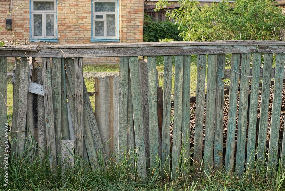 Sticker old gray wooden fence wall with broken boards overgrown with green vegetation on a rural street