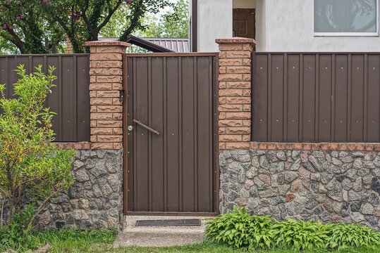 one brown metal door on the wall of a fence made of bricks and iron on a gray stone foundation in a street in green vegetation