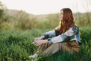 Fototapeta premium A young beautiful woman sits on the green grass in the park and looks out into the setting sun