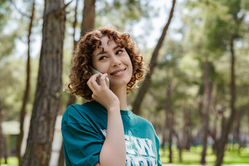 Side view of young woman wearing green t-shirt standing on city park, outdoors talking on mobile phone with friends or boyfriend with smiles.