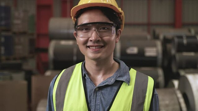 Portrait of happy professional Asian man worker wearing safety goggles and helmet standing smiling and looking at camera in the factory. Close up of handsome young male industry engineer in warehouse.