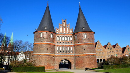 Fototapeta na wymiar Lübeck Holstentor mit zwei Spitztürmen neben Salzspeichern und Grünanlage unter blauem Himmel 