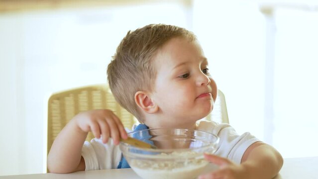 Baby eats cereal porridge. Bboy is holding spoon in his hand. Child sits at table and eats porridge.Family breakfast in kitchen. Feeding child at table.Child proper nutrition.Breakfast in kindergarten