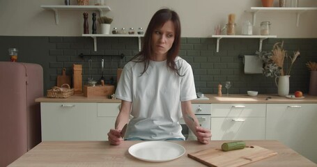 Woman sitting at table, looking sad and bored with diet not wanting to eat slice of cucumber. Healthy nutrition and dieting eating